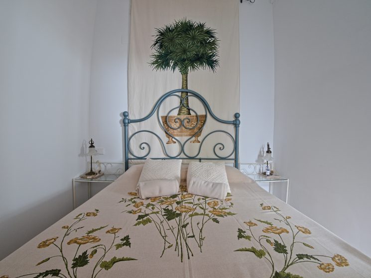 Bedroom at Casa Mayolica with blue wrought-iron headboard, palm tree tapestry and floral bedspread in Manises, Valencia