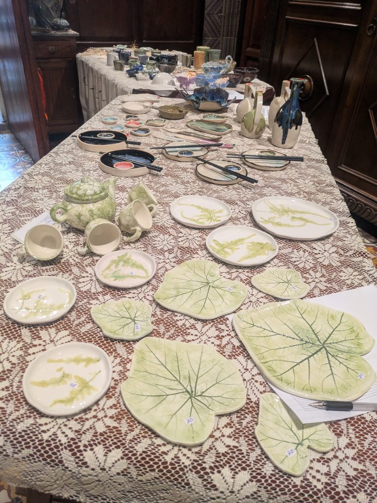 Table with a white lace cloth displaying handmade ceramics for sale at the Casa Mayolica market: leaf-shaped plates, a green-motif teapot with cups, bowls and small dishes with price tags