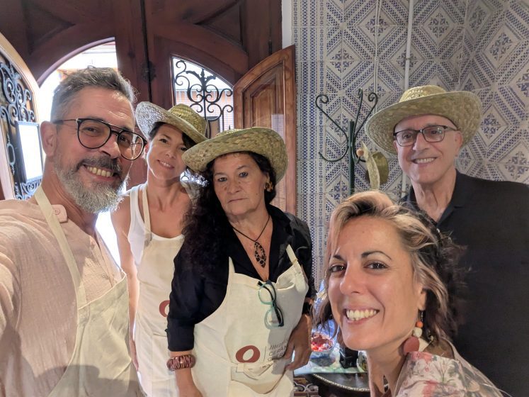 Five members of the Casa Mayolica team in straw hats and white aprons taking a selfie in front of blue and white tiled walls, the patio doorway open behind them, on the morning of Ceràmica Oberta 2026